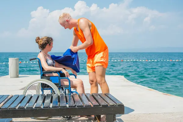 Plaj cankurtaranı engelli birine yardım ediyor. Deniz kenarında güneşli bir günde tekerlekli sandalyede oturan genç bir kadın için mavi havlu tutuyor..