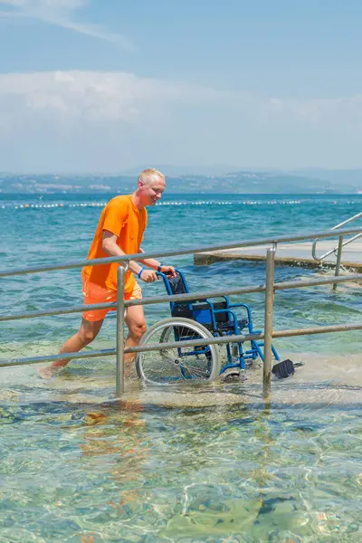 Gülümseyen cankurtaran, denizden gelen boş bir tekerlekli sandalyeyi sahil rampası boyunca itiyor, ulaşılabilir bir turizm hizmeti, bir çevre, ve bir yaz durağı, ön manzara..