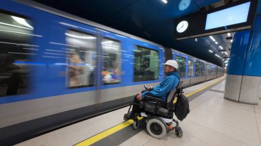Engelli bir adam, tekerlekli sandalyesinde beyaz bir şapka ve sırt çantası var, bağımsız seyahatin tadını çıkarıyor ve metro treninin gelmesini bekliyor..