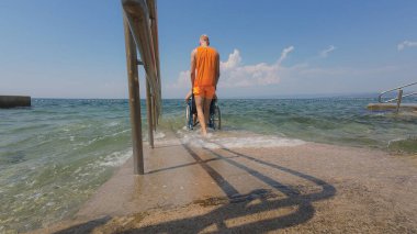 Yardımsever erkek asistan dalgalı denizde ferahlatıcı bir banyodan sonra ulaşılabilir sahil rampası boyunca tekerlekli sandalyedeki bir kadına dikkatlice rehberlik etti..