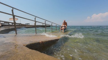 Engelli bir kadın, asistanının yardımıyla deniz suyundan çıkıyor, erişilebilir tekerlekli sandalye plaj rampasından geçiyor..