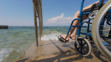 Erkek asistan, engelli bir kadını, yaz tatilinde dinlenmek için, dalgalı deniz kenarındaki ulaşılabilir sahil rampasına itiyor..