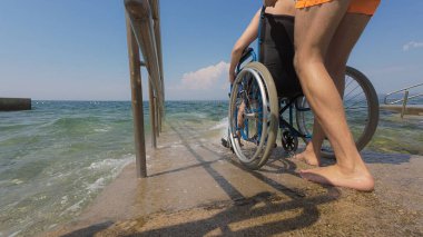 Can kurtaran engelli bir kadının halka açık bir plajdaki erişilebilir tekerlekli sandalye rampasından suya girmesine yardım ediyor..