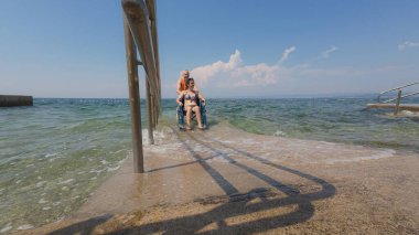 Engelli bir kadın, asistanının yardımıyla deniz suyundan çıkıyor, erişilebilir tekerlekli sandalye plaj rampasından geçiyor..