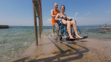 Denize tam erişimi olan engelli bir kadın sudan sahile kadar cankurtaran yardımcılarıyla ulaşılabilir bir rampadan geçiyor..