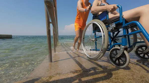 Can kurtaran engelli bir kadının halka açık bir plajdaki erişilebilir tekerlekli sandalye rampasından suya girmesine yardım ediyor..