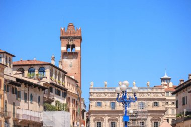 Ziyaretçiler, tarihi binalarla çevrili Verona, İtalya 'da canlı bir meydanda geziniyorlar. Güneş parıldıyor, saat kulesinin ve etrafındaki cephelerin mimari detaylarını aydınlatıyor..