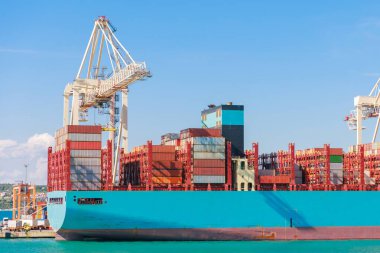 Kalabalık bir limana yanaşan büyük bir konteyner gemisi renkli kargo konteynırlarını boşaltıyor. Açık mavi gökyüzü ve sakin sular deniz aktivitesini vurgular.