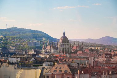 Budapeşte 'nin sembolik Parlamento Binası, şehir çatıları ve yeşil tepelerle çevrili, parlak mavi gökyüzüne karşı belirgin bir şekilde duruyor ve güzel bir şehir manzarası sergiliyor..