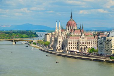 Budapeşte Parlamentosu binası Tuna Nehri boyunca berrak bir gökyüzünün altında görkemli bir şekilde duruyor. Tekneler denizde dolaşırken, yeşil tepeler manzaralı bir zemin oluşturuyor..