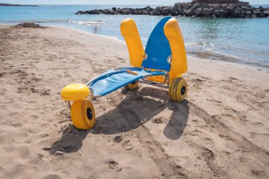 Parlak renkli plaj tekerlekli sandalyesi suyun yakınındaki sıcak kumun üzerine park edilmiştir. Dalgalar kıyı şeridinde yavaşça dolaşırken, bazı insanlar arka planda plajın tadını çıkarıyor..