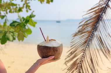 Tayland 'da pitoresk bir plajda bir el taze hindistan cevizi içeceği tutuyor. Kristal berrak su ve uzak yelkenliler dinlenmek için mükemmel bir tropikal atmosfer yaratır..