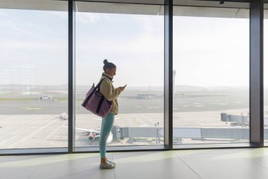 Bir yolcu bir havaalanının büyük penceresinin yanında duruyor, bir uçuş beklerken telefonlarını kontrol ediyor. Arka planda pist ve kapılar görülebiliyor, dışarıda uçak park halinde..