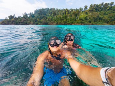Tayland açıklarındaki çarpıcı turkuaz sularda şnorkelle yüzen iki birey, yemyeşil sularla çevrili ve güneşli bir günde canlı deniz yaşamının tadını çıkarıyorlar..