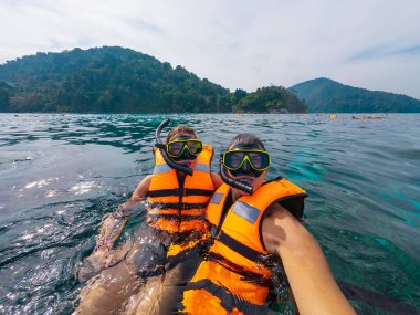 İki arkadaş, canlı deniz yaşamını keşfederken parlak turuncu can yelekleri giyip Tayland 'da şnorkelle dalmayı seviyor. Yeşil yemyeşil dağlar arka planı çerçeveleyerek tropikal atmosferi güçlendirir..