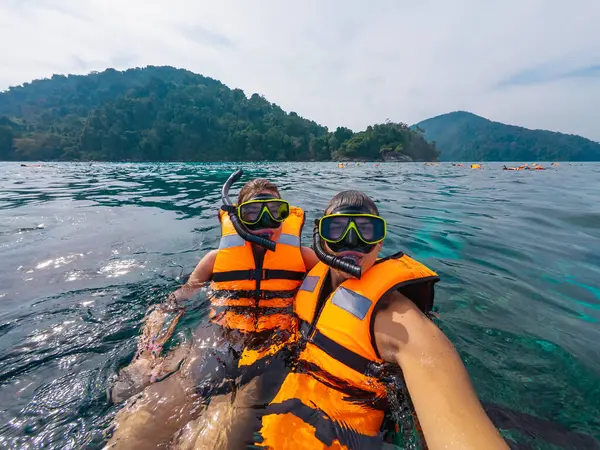 İki arkadaş, canlı deniz yaşamını keşfederken parlak turuncu can yelekleri giyip Tayland 'da şnorkelle dalmayı seviyor. Yeşil yemyeşil dağlar arka planı çerçeveleyerek tropikal atmosferi güçlendirir..