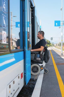 Erişilebilir tren yolculuğu kullanan tekerlekli sandalye kullanıcısı bir istasyondaki trene yaklaşır. Kullanıcı, arka planda açık işaret ile tren kapısı açılır açılmaz binmeye hazırlanır.