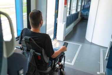 Erişilebilir tren yolculuğu kullanan tekerlekli sandalye kullanıcısı, telefonlarını kontrol ederken modern bir trende rahat bir yolculuğun keyfini çıkarıyor. İç kısımda tüm yolcular için tasarlanmış geniş koltuklar bulunmaktadır..
