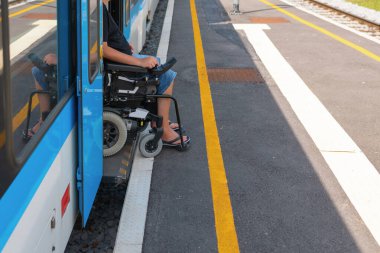 Erişilebilir tren yolculuğu kullanan tekerlekli sandalye kullanıcısı trenin yanındaki platformda bekliyor. Çevrede açık çizgiler ve herkes için güvenli erişim sağlayan iyi korunmuş bir yol var..