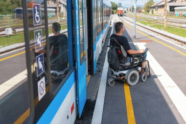 Erişilebilir tren yolculuğu kullanan tekerlekli sandalye kullanıcısı bir platformda bekliyor, gelen trene binmeye hazır. Çevrede açık yollar ve erişilebilirlik sembolleri var, içeriği vurguluyor.