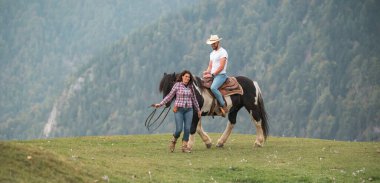 Bir adam çimenli bir tepede siyah beyaz bir ata binerken, bir kadın dizginleri elinde tutarak onların yanında yürüyor. Arka planda dağlar görünür..