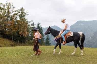 Kovboy kız eğitmeni, dizginleri elinde tutuyor, çiftlik biniciliği okulunda ata binen genç bir sürücüyü izliyor ve ona yardım ediyor.