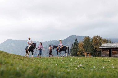 Dağlardaki yeşil çimenli bir arazide batı kıyafeti giymiş bir grup insan ata biniyor. Küçük ahşap bir binanın yanında bir köpek duruyor..
