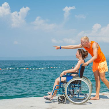 Engelli gülümseyen bir kadın ve asistanı tekerlekli sandalyesinin hareket etmesine yardım ediyor, sahilde ve deniz ufkunda eğleniyor..
