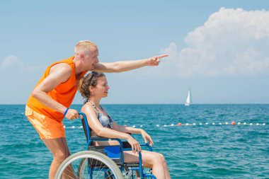 Engelli gülümseyen bir kadın ve asistanı tekerlekli sandalyesinin hareket etmesine yardım ediyor, sahilde ve deniz ufkunda eğleniyor..
