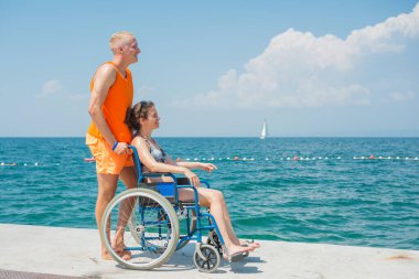 Engelli gülümseyen bir kadın ve asistanı tekerlekli sandalyesinin hareket etmesine yardım ediyor, sahilde ve deniz ufkunda eğleniyor..