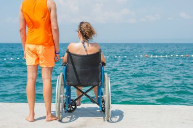 Engelli gülümseyen bir kadın ve asistanı tekerlekli sandalyesinin hareket etmesine yardım ediyor, sahilde ve deniz ufkunda eğleniyor..