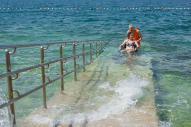 Rahatlamış, engelli bir kadın, bir asistanın yardımıyla deniz suyundan çıkıyor, erişilebilir tekerlekli sandalye plaj rampasından geçiyor..