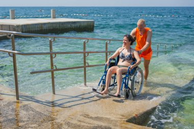 Denize tam erişimi olan engelli bir kadın sudan sahile kadar cankurtaran yardımcılarıyla ulaşılabilir bir rampadan geçiyor..