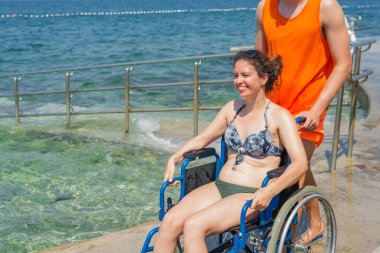 Yardımsever bir erkek asistan, mavi denize doğru uzanan ulaşılabilir sahil rampası boyunca, tekerlekli sandalyeli bir kadını ittiriyor..