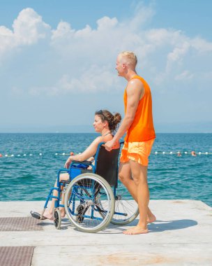Birlikte, engelli gülümseyen bir kadın, tekerlekli sandalye kullanıcısı ve sahil görevlisi asistanı yaz güneşinin altındaki güzel deniz manzarasının tadını çıkarıyorlar..