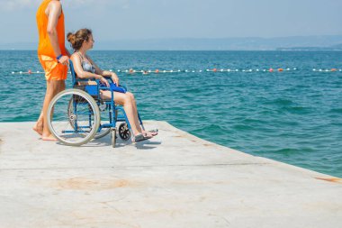 Birlikte, engelli gülümseyen bir kadın, tekerlekli sandalye kullanıcısı ve sahil görevlisi asistanı yaz güneşinin altındaki güzel deniz manzarasının tadını çıkarıyorlar..