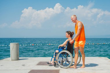Engelli gülümseyen bir kadın ve asistanı, tekerlekli sandalyesinin hareket etmesine yardım eden bir plaj görevlisi, güneşli bir yaz gününde güzel bir deniz manzarasının keyfini çıkarıyorlar..