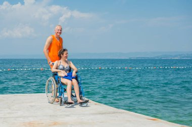 Yardımsever bir erkek asistan, mavi denize doğru uzanan ulaşılabilir sahil rampası boyunca, tekerlekli sandalyeli bir kadını ittiriyor..