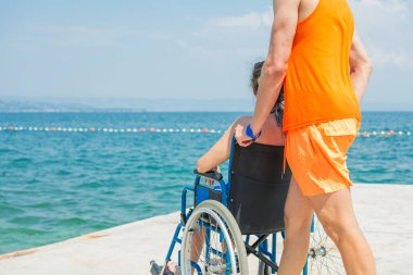 Yardımsever bir erkek asistan, mavi denize doğru uzanan ulaşılabilir sahil rampası boyunca, tekerlekli sandalyeli bir kadını ittiriyor..