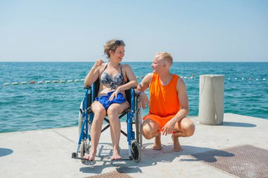 Engelli gülümseyen bir kadın ve asistanı, tekerlekli sandalyesinin hareket etmesine yardım eden bir plaj görevlisi, güneşli bir yaz gününde güzel bir deniz manzarasının keyfini çıkarıyorlar..