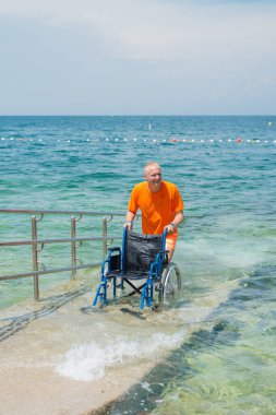 Gülümseyen cankurtaran, arka planda güzel bir deniz manzarası olan bir yaz turistik bölgesinde ulaşılabilir bir sahil rampası boyunca boş bir su tekerlekli sandalyesini yuvarlıyor..