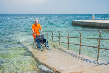 Gülümseyen cankurtaran, arka planda güzel bir deniz manzarası olan bir yaz turistik bölgesinde ulaşılabilir bir sahil rampası boyunca boş bir su tekerlekli sandalyesini yuvarlıyor..