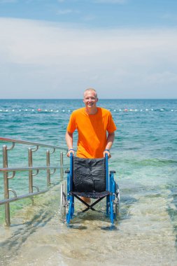 Gülümseyen cankurtaran, arka planda güzel bir deniz manzarası olan bir yaz turistik bölgesinde ulaşılabilir bir sahil rampası boyunca boş bir su tekerlekli sandalyesini yuvarlıyor..