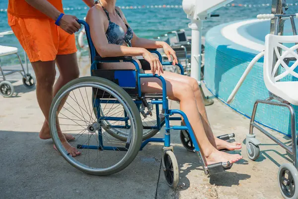 Deniz oteli cankurtaranı engelli bir kadına yardım ediyor erişilebilir hizmetler ve tesisler, havuz sandalyesi kaldırma, ve tekerlekli sandalye, tatil köyünde..