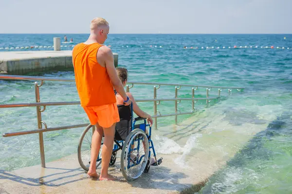 Engelli bir kadın, deniz suyuna bir cankurtaran yardımcısının yardımıyla erişilebilir tekerlekli sandalye plaj rampasından giriyor..
