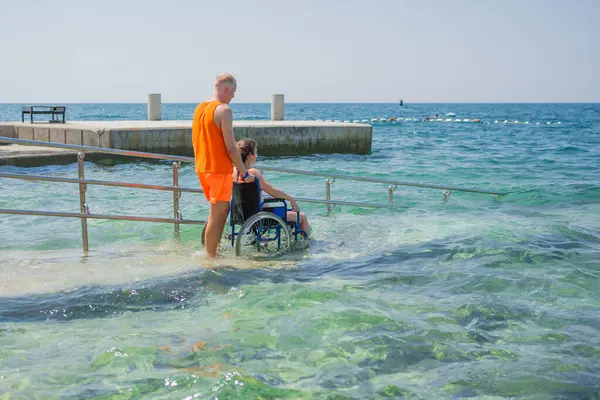 Can kurtaran engelli bir kadının halka açık bir plajdaki erişilebilir tekerlekli sandalye rampasından suya girmesine yardım ederek, denizin ve dikiz aynasının tadını çıkarmasına izin veriyor..