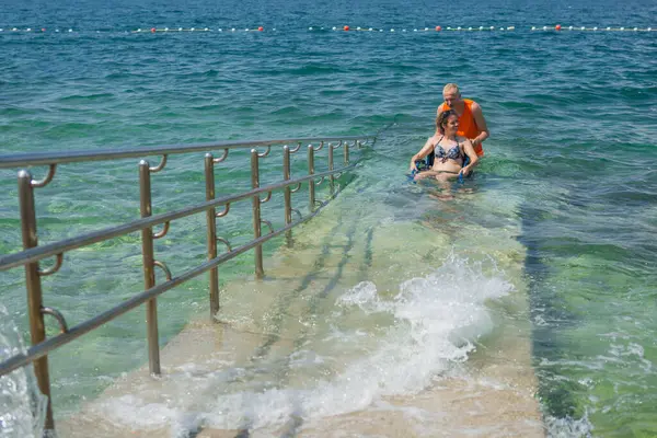 Rahatlamış, engelli bir kadın, bir asistanın yardımıyla deniz suyundan çıkıyor, erişilebilir tekerlekli sandalye plaj rampasından geçiyor..