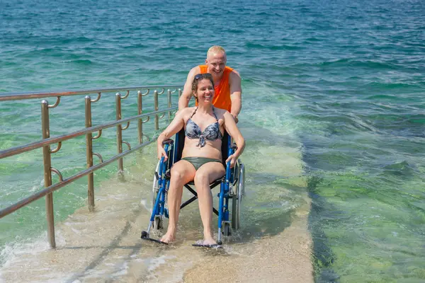Yaz tatilinde gülümseyen genç bir çift, tekerlekli sandalyede karısına yardım eden bir adam, deniz manzaralı ferahlatıcı bir banyodan sonra ulaşılabilir sahil rampasında onu iten bir adam..