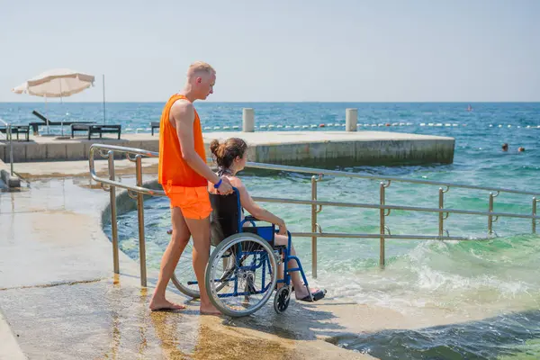 Can kurtaran engelli bir kadının halka açık bir plajdaki erişilebilir tekerlekli sandalye rampasından suya girmesine yardım ederek, denizin ve dikiz aynasının tadını çıkarmasına izin veriyor..