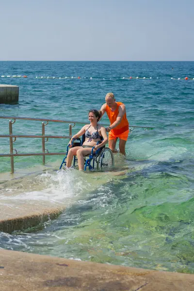 Rahatlamış, engelli bir kadın, bir asistanın yardımıyla deniz suyundan çıkıyor, erişilebilir tekerlekli sandalye plaj rampasından geçiyor..